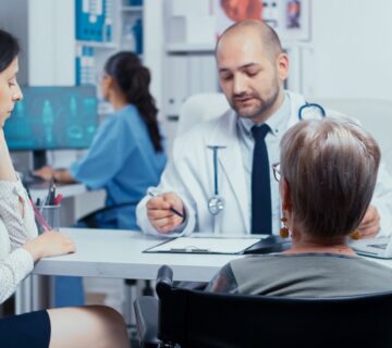 A doctor converses with a patient in a wheelchair, discussing health concerns in a clinical setting.