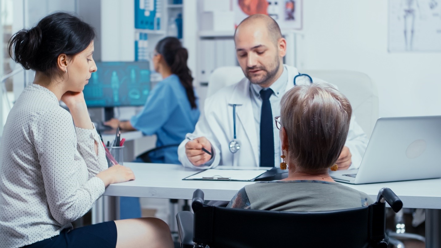 A doctor converses with a patient in a wheelchair, discussing health concerns in a clinical setting.