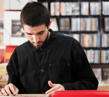 A man examines a vinyl record while browsing in a record store, surrounded by shelves of albums.
