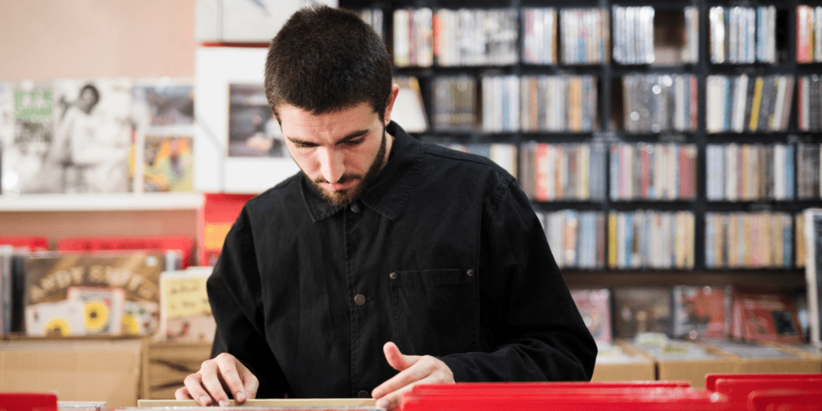 A man examines a vinyl record while browsing in a record store, surrounded by shelves of albums.